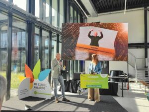 Een man spreekt op het podium tijdens de uitreiking van De Cirkel Awards in een lichte ruimte met hoge ramen. Een vrouw ontvangt een symbolische cheque. Op het grote scherm achter hen staat een afbeelding van een juichende man met de tekst "Eromesmarko". De sfeer is feestelijk en professioneel.