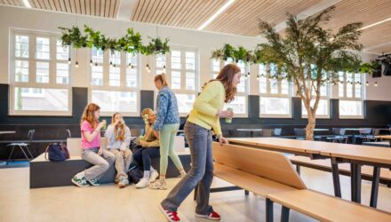 Een groep leerlingen in een leerplein-achtige ruimte. Op de voorgrond zit een leerling aan een tafelbank met opklapbare rugleuning. De rest van de groep zit op een zitelement, ontspannen met een drankje. De ruimte is licht en groen ingericht, met planten aan het plafond en een boom in het midden.