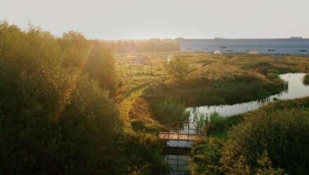Een ochtendzon schijnt over de circulaire proeftuin Greenfield, met waterpartijen, groene paden en open houten structuren. Op de achtergrond is het gebouw van Fair Furniture Group zichtbaar.